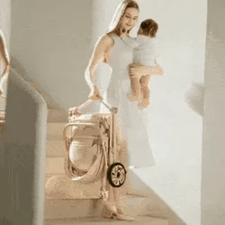 Woman in a white dress holding a baby and walking with a baby buggy in a bright room.